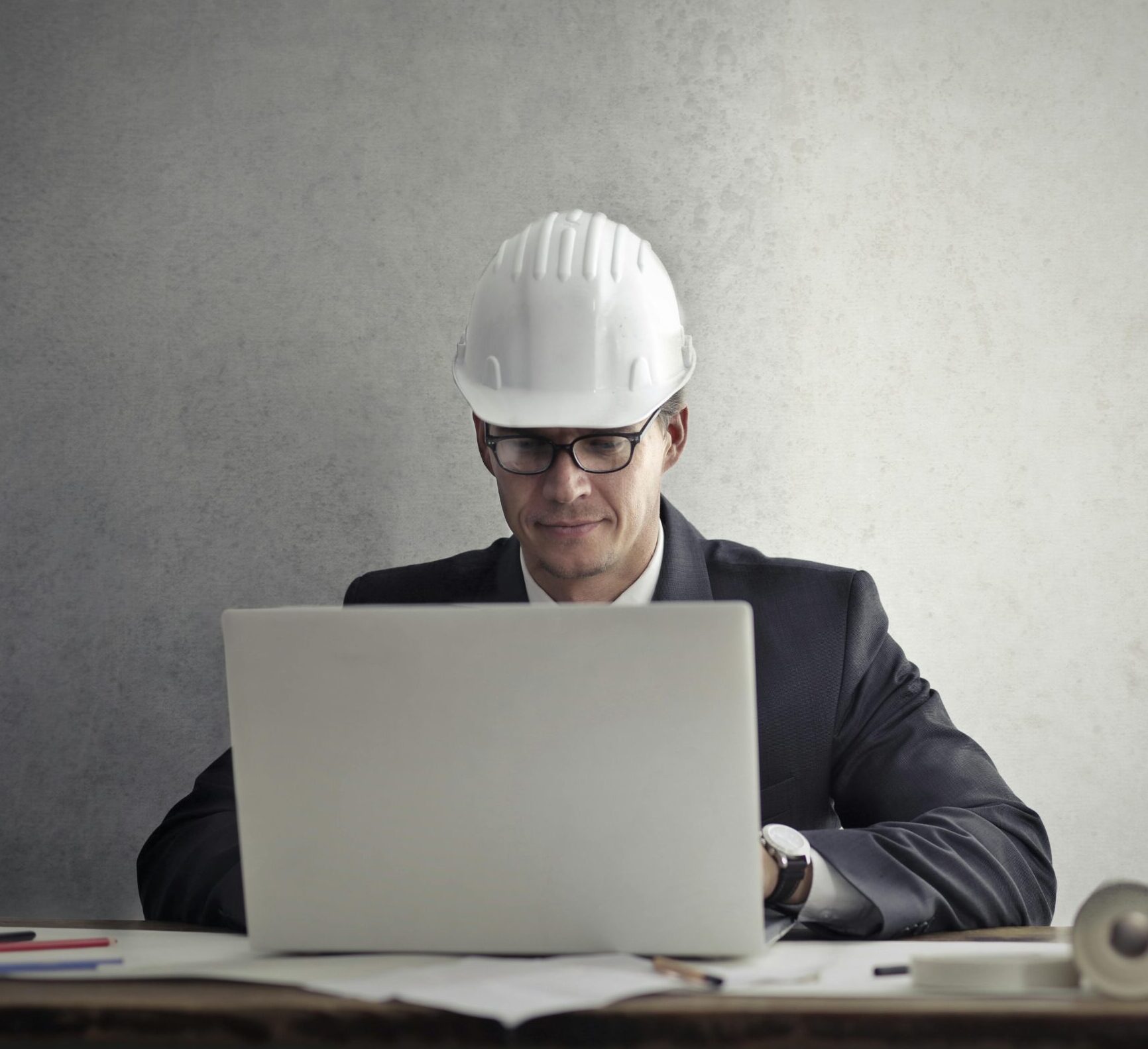 Professional engineer wearing hardhat working on construction plans at desk with laptop in office.